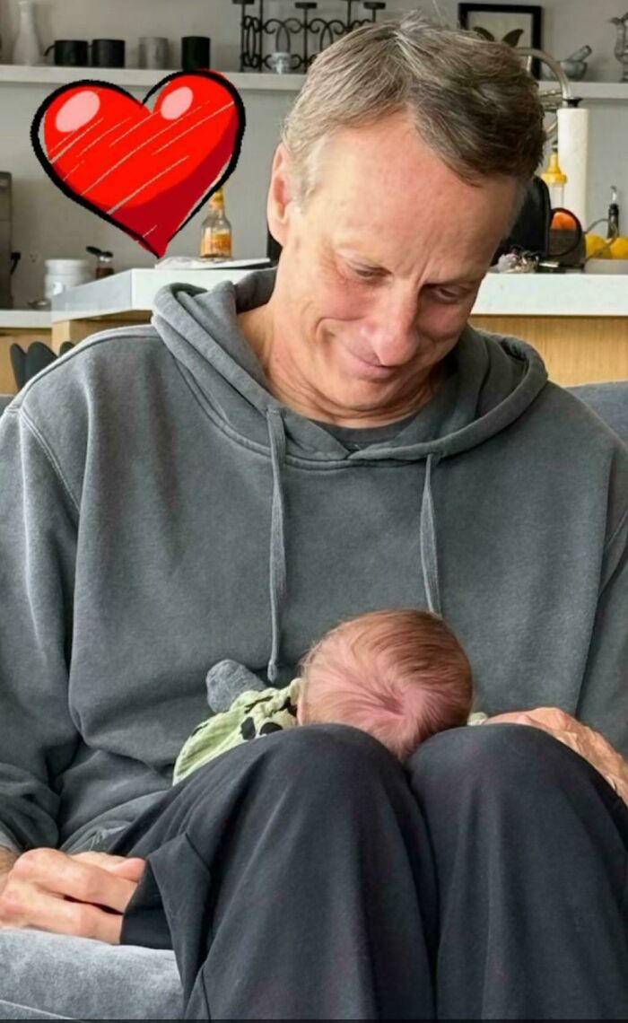 Man wearing gray hoodie holding a newborn baby on his lap with a red heart graphic in a cozy home setting.