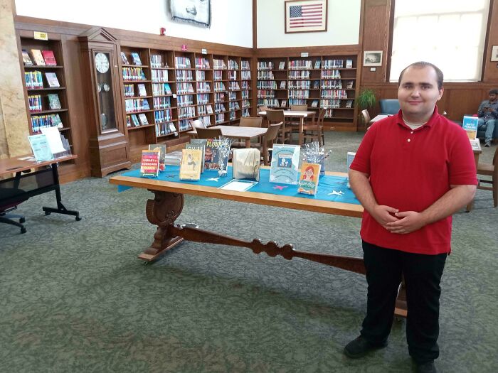 Young man smiling in a library with books on a table, creating a heartwarming and uplifting atmosphere.