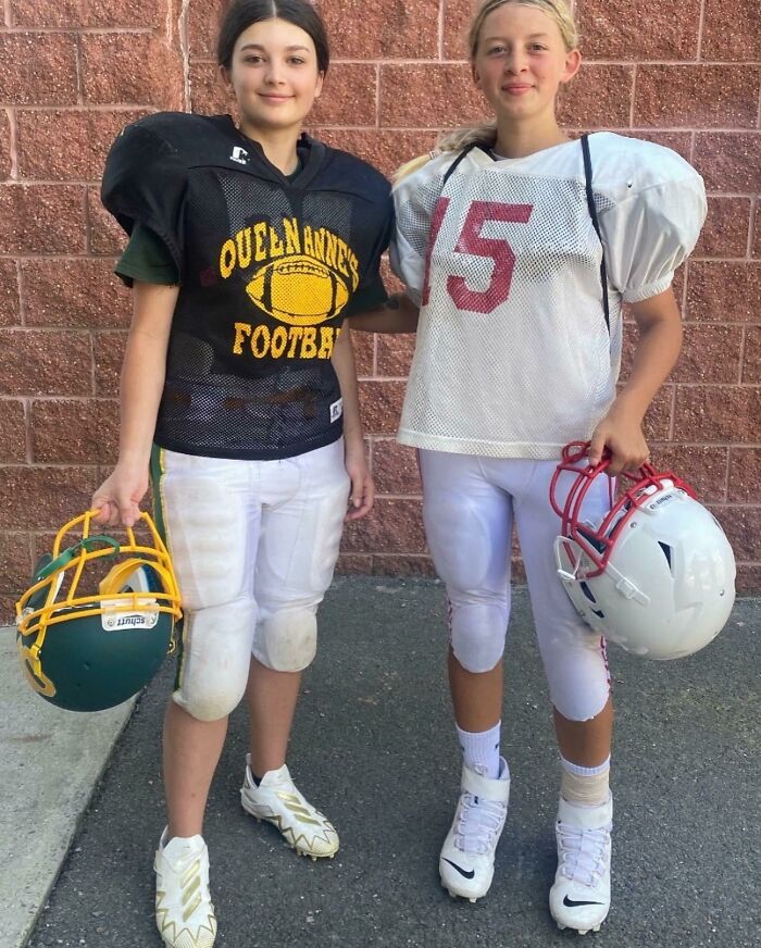 Two young girls in football gear smiling and holding helmets, capturing a heartwarming moment of friendship and teamwork.
