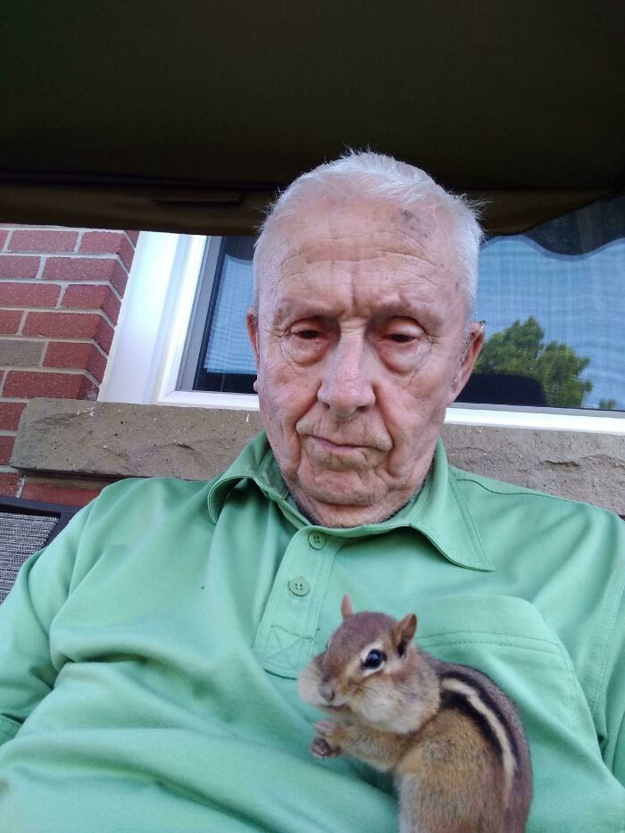 Elderly man in a green shirt sitting outside with a chipmunk on his chest, capturing a heartwarming moment.