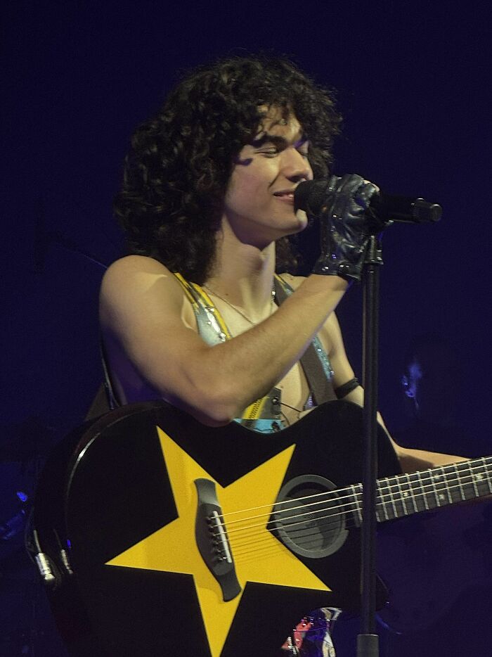 Conan Gray singing on stage playing a black guitar with a large yellow star during a live performance.