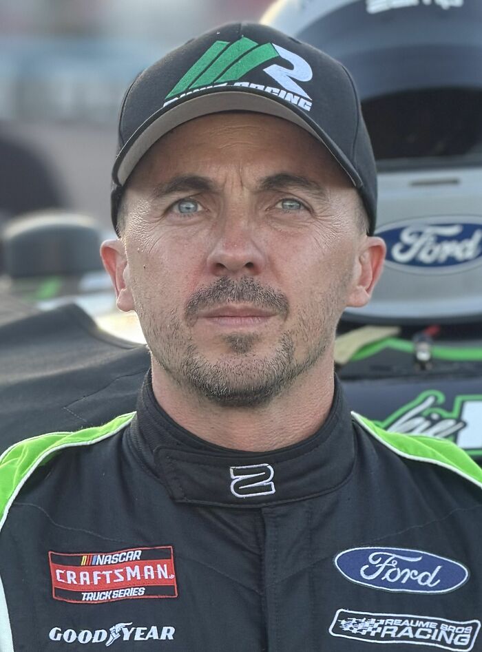 Frankie Muniz wearing racing suit and cap with Ford and NASCAR Craftsman Truck Series logos at a race event.