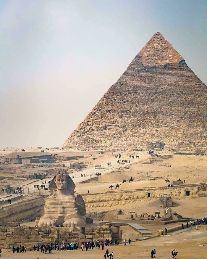 Great Pyramid and Sphinx in Ancient Egypt with tourists and camels in the desert landscape under clear sky.