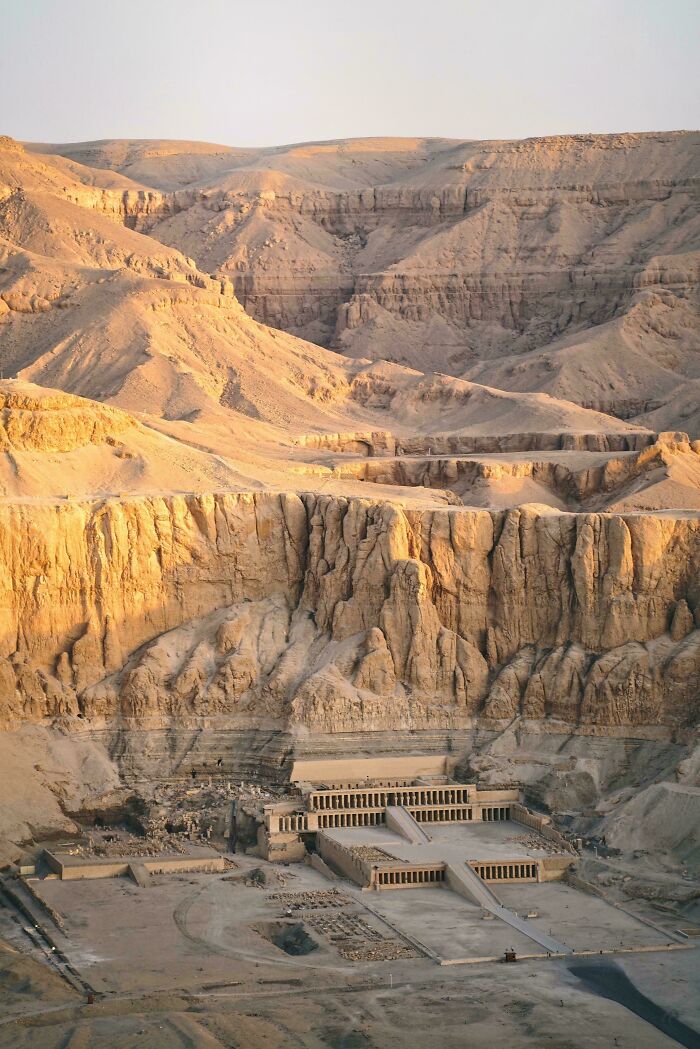 Ancient Egypt archaeological site carved into desert cliffs with temple structures at the base in warm sunlight.