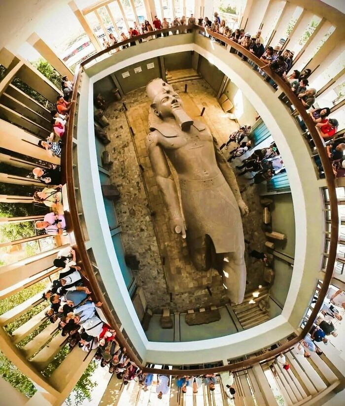 Large ancient Egyptian statue of a pharaoh viewed from above, surrounded by tourists in a museum setting.
