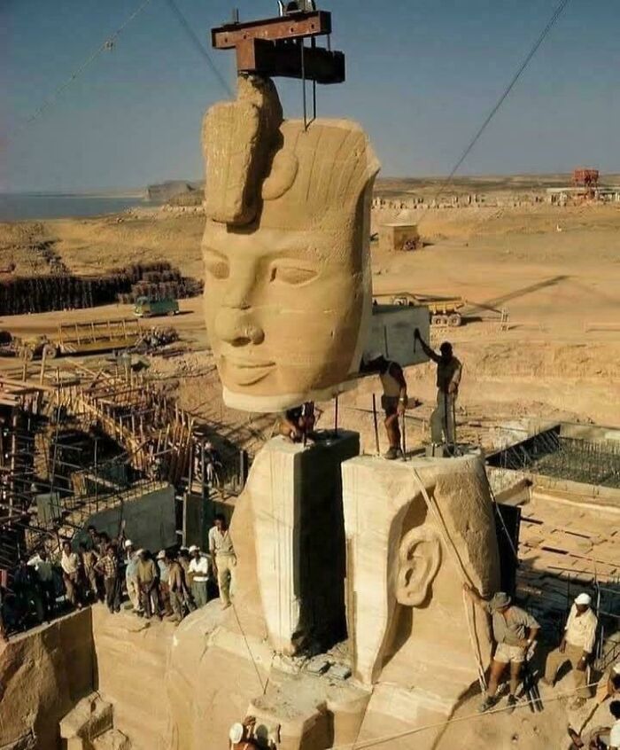 Large ancient Egyptian statue head being lifted by crane during excavation with workers and desert landscape in background.