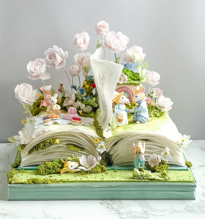 Intricately crafted cake shaped like an open book with mouse figurines, flowers, and detailed edible scenery.