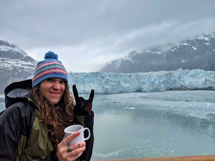 Woman in winter gear holding a cup and making a hand gesture near a glacier, showcasing unique job moments.