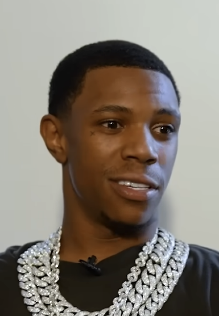 A Boogie wit da Hoodie wearing multiple diamond chains and a black shirt, speaking in an interview setting.