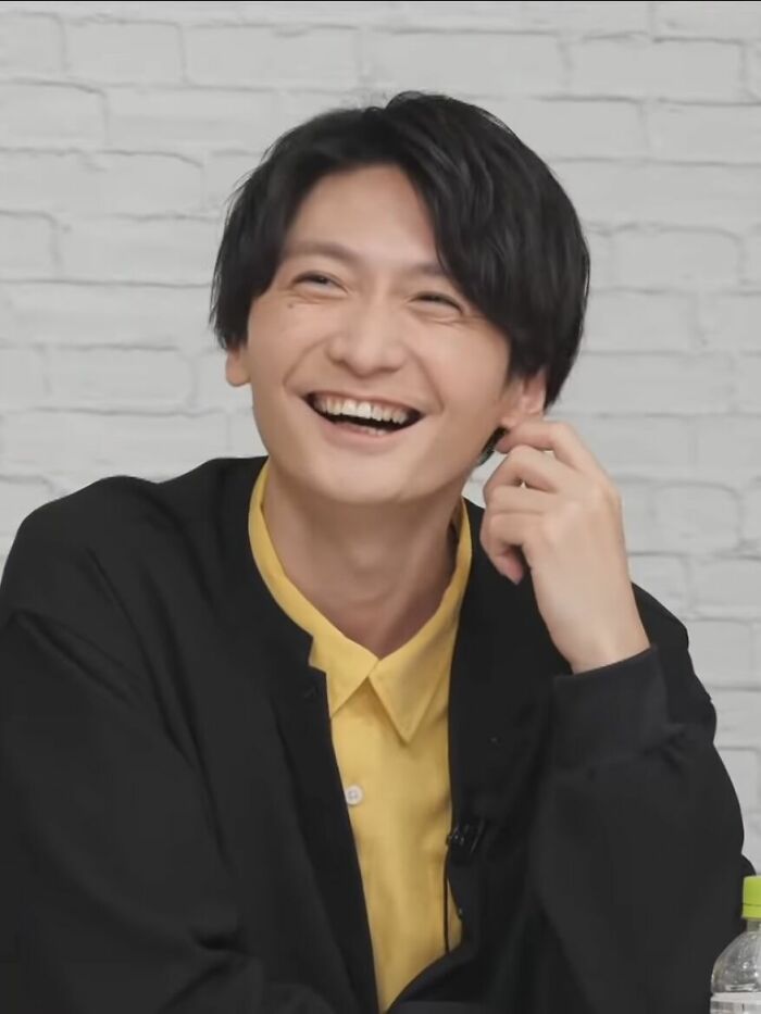 Nobunaga Shimazaki smiling during an interview, wearing a yellow shirt and black jacket against a light brick wall background.