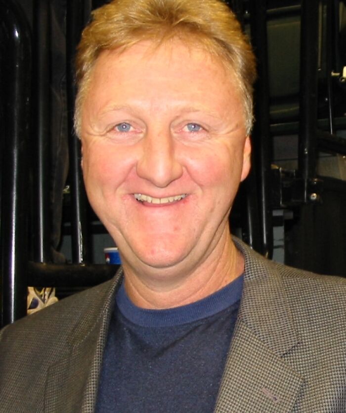 Larry Bird smiling in a casual blazer and shirt, highlighting key moments from his bio and career highlights.