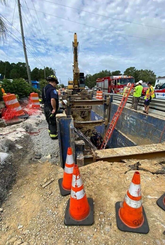 Construction workers ignoring safety protocol while working near a deep open trench with emergency vehicles nearby.