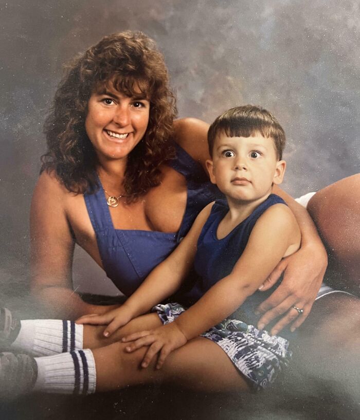 Woman smiling in blue outfit holding wide-eyed child in sleeveless shirt in a hilariously awkward childhood photo.