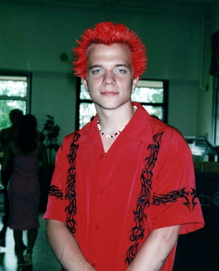 Teen with bright red spiked hair wearing a red shirt with black designs, a necklace, posing for an awkward childhood photo.