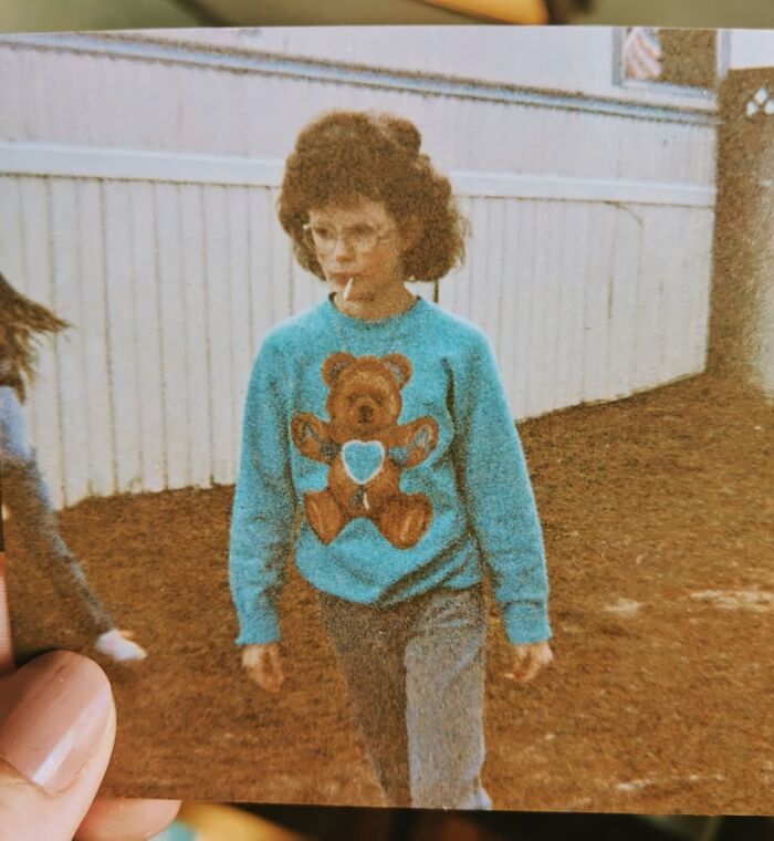 Child wearing teddy bear sweater and glasses in a vintage awkward childhood photo outdoors by a fence.