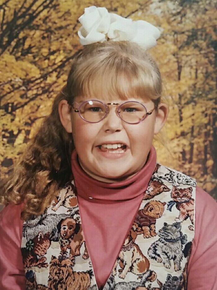 Awkward childhood photo of a girl with glasses, a big hair bow, and a vest covered in dog and cat patterns.