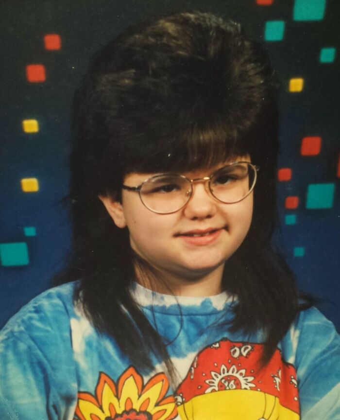 Teen with large 80s hairstyle and glasses wearing a colorful tie-dye shirt in a hilariously awkward childhood photo.