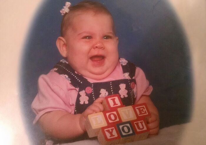Chubby baby girl wearing pink holding wooden blocks spelling I love you in a hilariously awkward childhood photo.