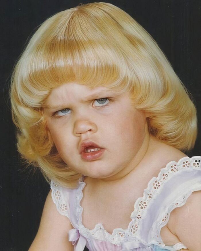 Toddler with blonde hair making a confused face in a hilariously awkward childhood photo with vintage style clothing.