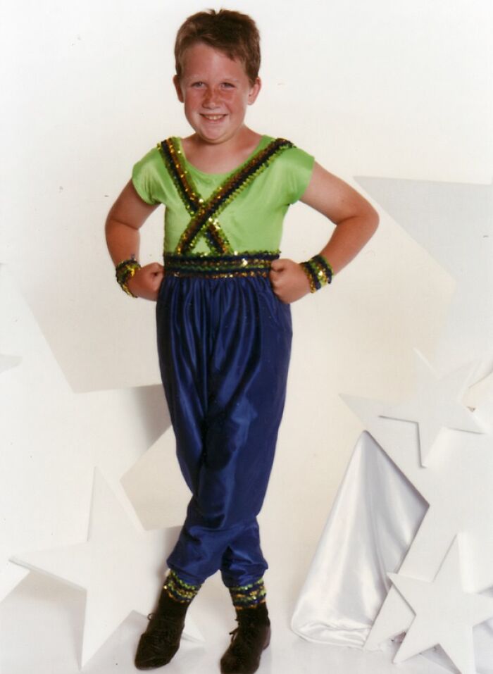 Smiling boy in a bright sequined costume posing awkwardly in front of a star-themed backdrop in a childhood photo.