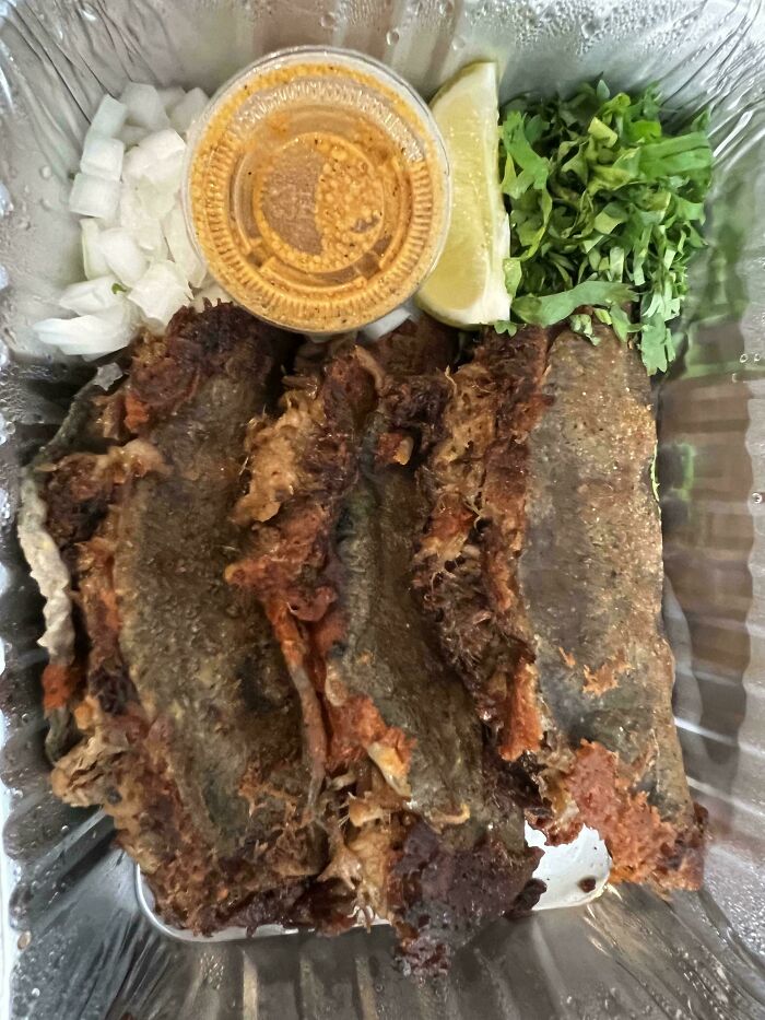 Three overcooked fish fillets in a takeout container with lemon wedge, chopped onions, cilantro, and sauce showing outrageous meals.