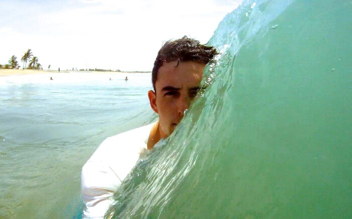 Man's face partially hidden by a wave captured in a funny and interesting weird pic taken at the perfect time at the beach.