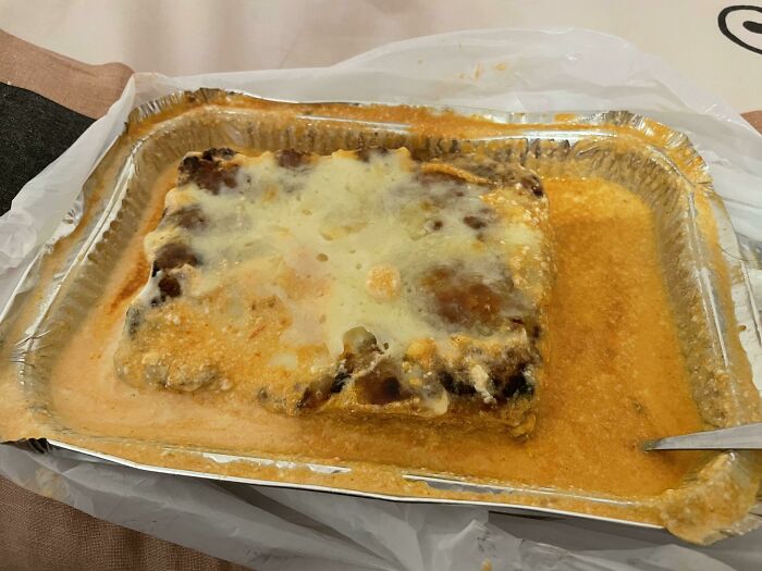 Overcooked lasagna in a foil tray covered in excess sauce, an example of outrageous meals that look nothing like menu photos.