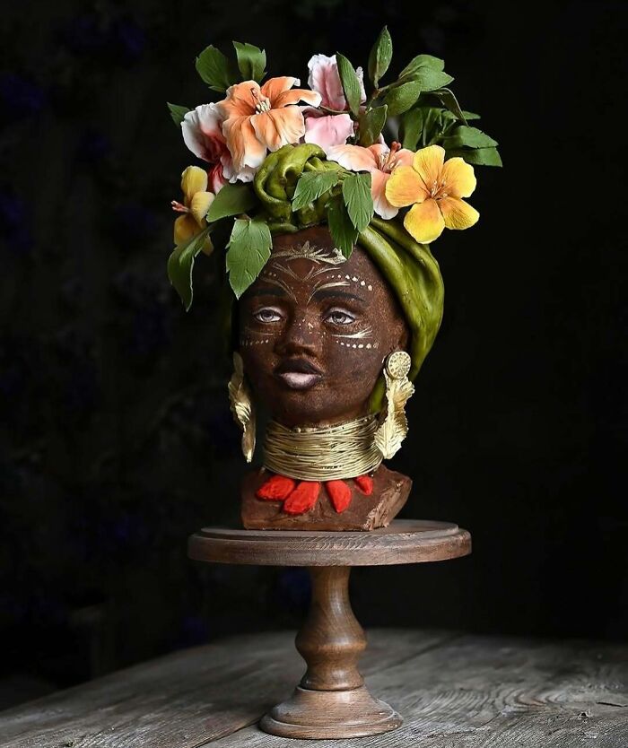 Masterfully crafted cake sculpted as a detailed human head with floral headpiece and intricate face designs on a wooden stand.