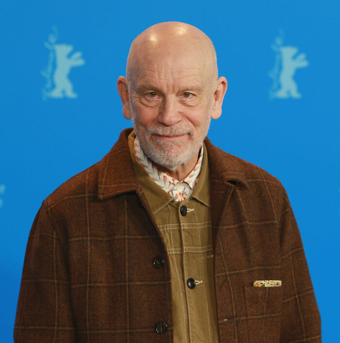 John Malkovich wearing a checked brown jacket and patterned shirt at a film festival photo call.