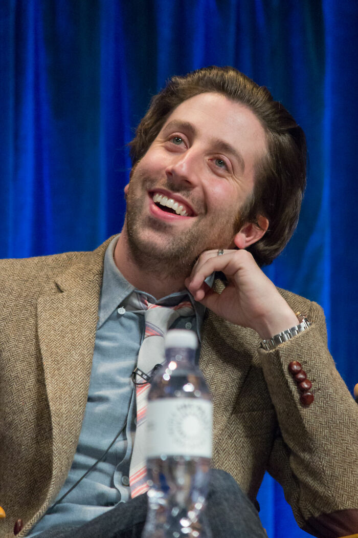 Simon Helberg smiling in a tan blazer and patterned tie during an interview with a blue curtain background.