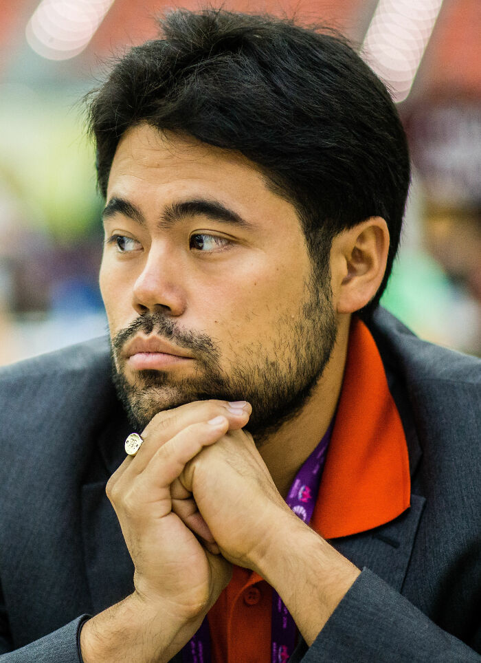 Hikaru Nakamura in a thoughtful pose during a chess event, wearing a dark jacket and an orange shirt.