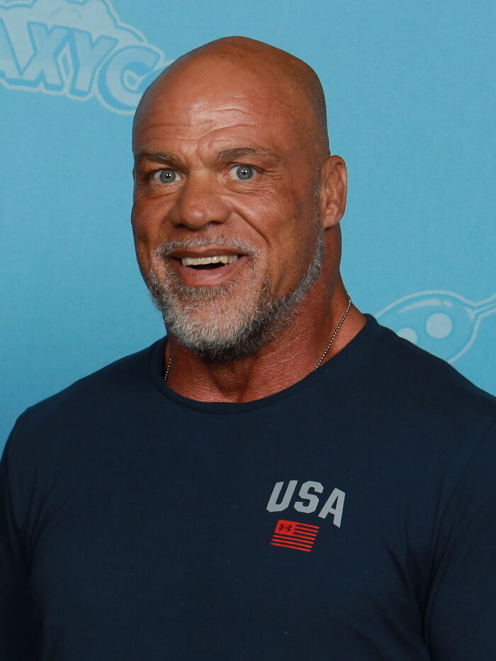 Kurt Angle smiling, wearing a navy USA shirt, showcasing his bio and career highlights in professional wrestling.