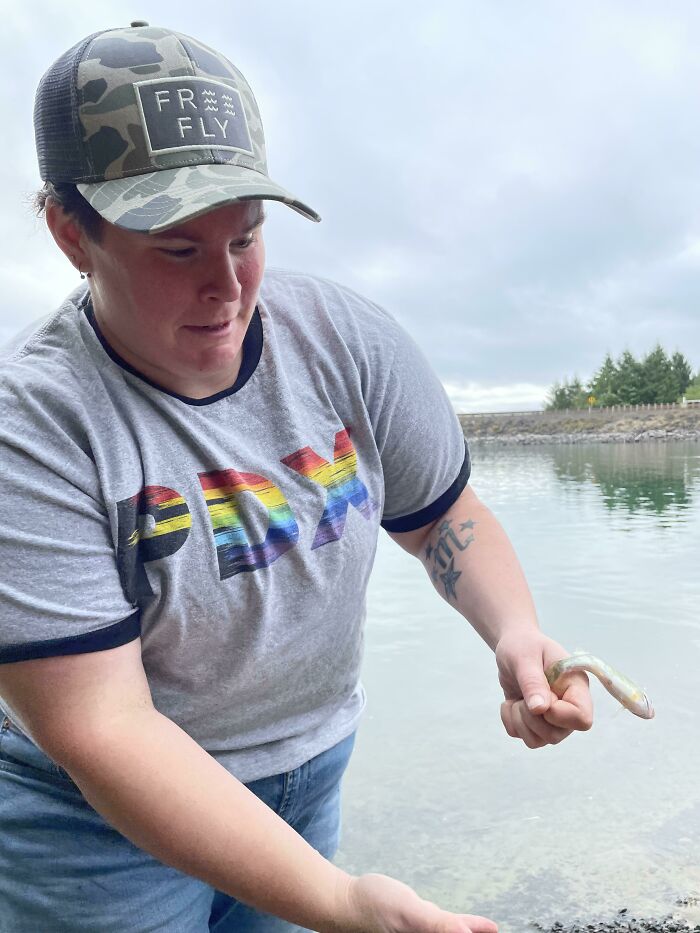 Person wearing a rainbow PDX shirt and camo hat holding a small fish by the water in a funny and interesting perfect timing pic.
