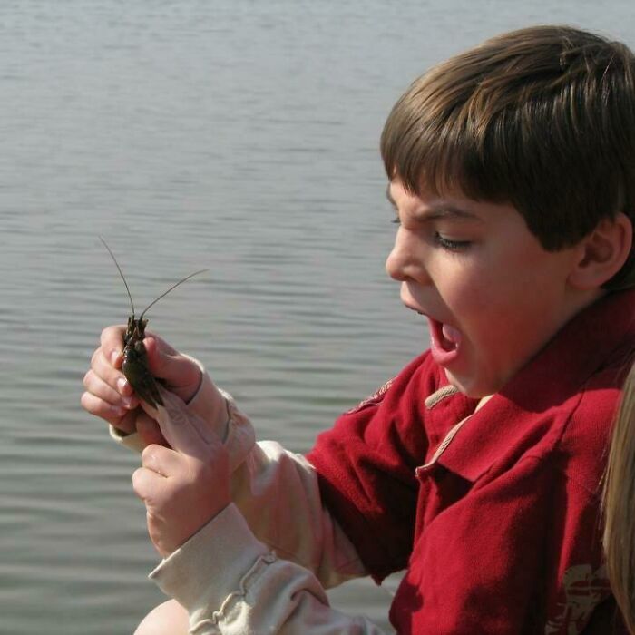 Young boy reacting with surprise while holding a crawfish near calm water in a funny perfect timing pic.
