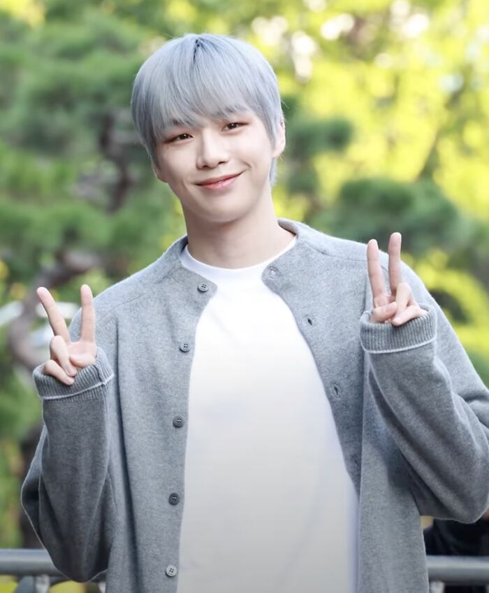 Kang Daniel smiling outdoors wearing a gray cardigan and white shirt, flashing peace signs with both hands in daylight.
