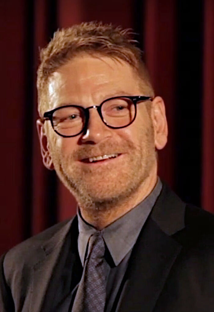 Kenneth Branagh wearing glasses and a suit, smiling during an interview focused on his bio and career highlights