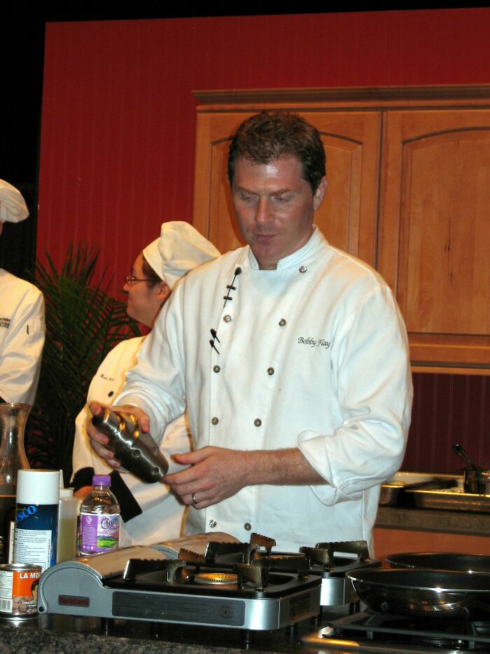 Bobby Flay wearing a chef’s coat, preparing food in a kitchen setting during a cooking demonstration.