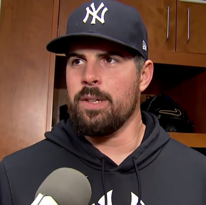 Carlos Rodón in a New York Yankees cap, speaking to reporters in a locker room setting after a game.