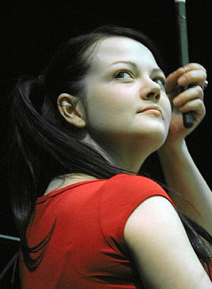 Meg White performing on stage, wearing a red top, with her hair tied back and holding drumsticks.