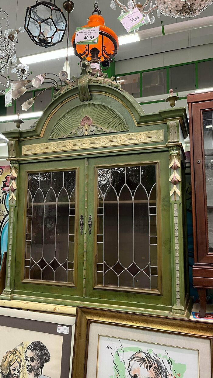 Vintage green wooden cabinet with decorative glass doors displayed in a secondhand store with lamps above.