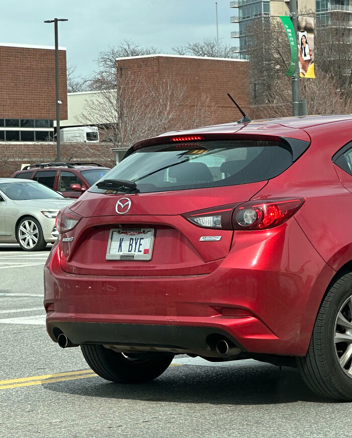 Red Mazda hatchback with a hilarious personalized license plate reading K BYE, parked in an urban area with other cars nearby.