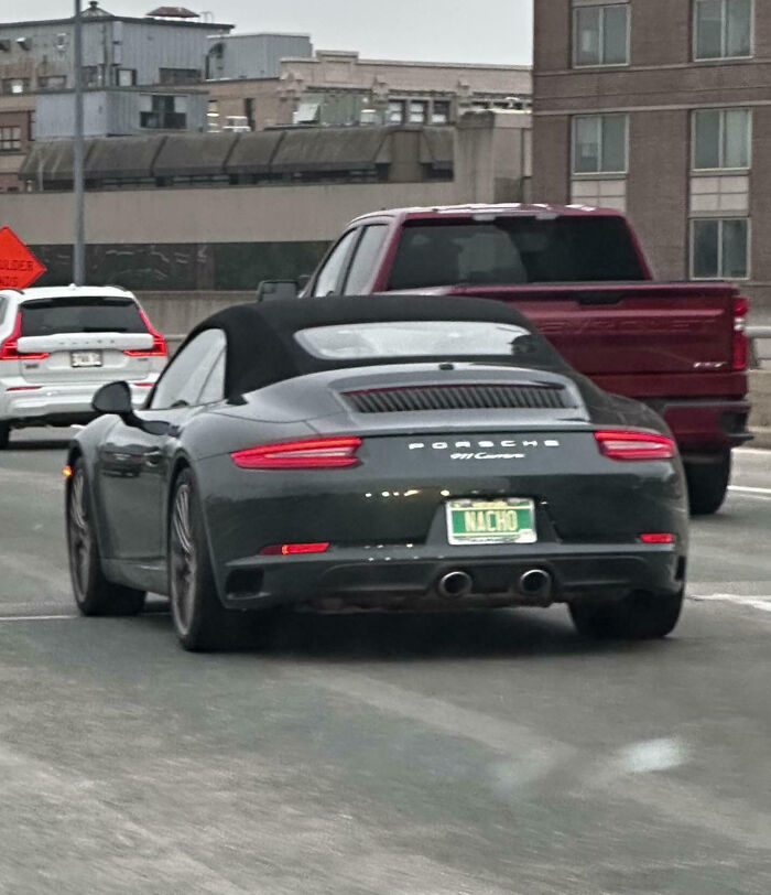 Black Porsche convertible with a hilarious personalized license plate reading NACHO on a city road.