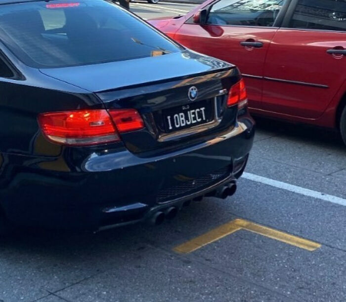 Black BMW with a personalized license plate reading I OBJECT parked next to a red car, showcasing funny personalized license plates.