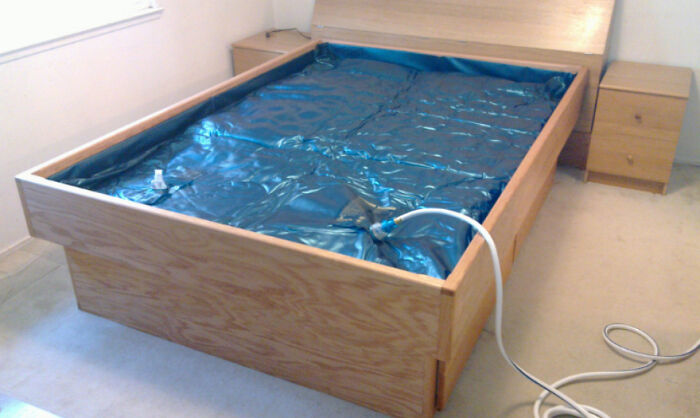 A wooden bed frame with a blue plastic-covered mattress area in a simple room with beige carpeting and two nightstands.