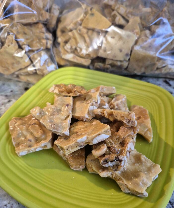 Broken pieces of peanut brittle on a green plate with a bag of brittle in the background, showing sugary texture.