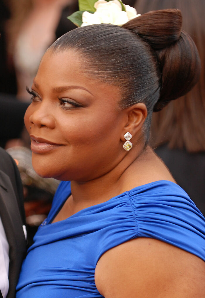 Mo'Nique smiling in a blue dress with a flower hair accessory, showcasing her bio and career highlights at an event