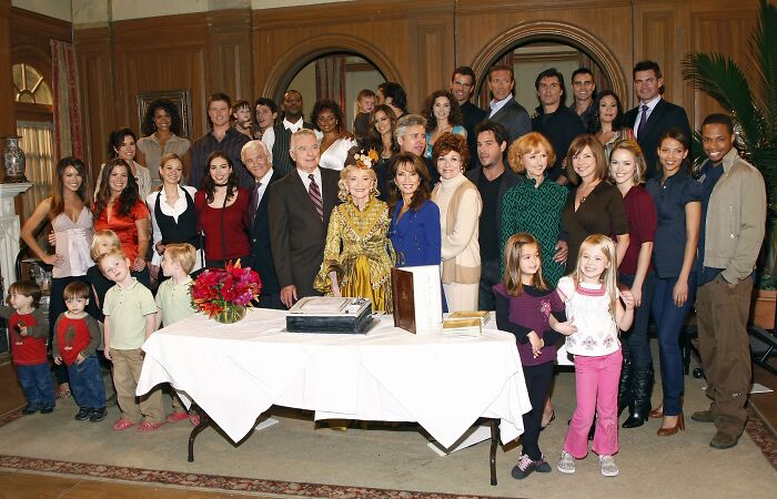 Large diverse cast of actors gathered in a room celebrating longest running TV shows with a cake on the table