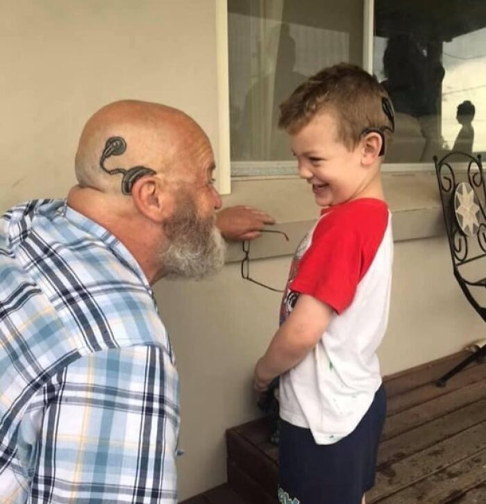 Man and boy smiling at each other showing matching wholesome tattoos behind their ears symbolizing meaningful family bond.