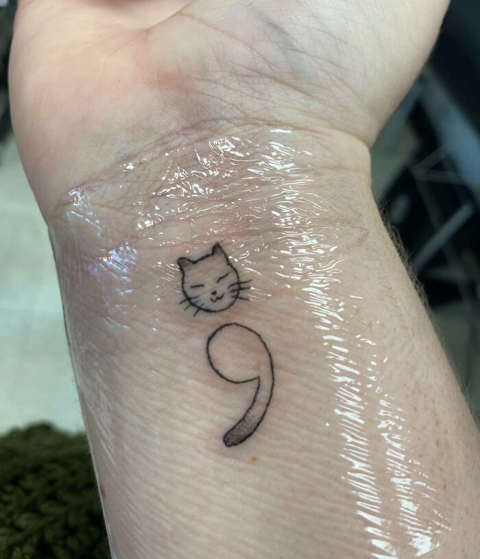 Minimalist cat and semicolon tattoo on wrist wrapped in protective film, showcasing wholesome tattoos with meaningful ink. Minimalist cat and semicolon tattoo on wrist wrapped in protective film, showcasing wholesome tattoos with meaningful ink.