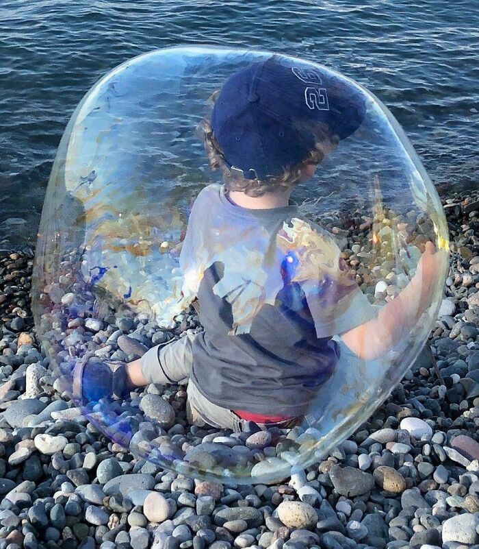 Child sitting on rocky shore inside a giant transparent bubble, a funny and interesting pic taken at the perfect time.